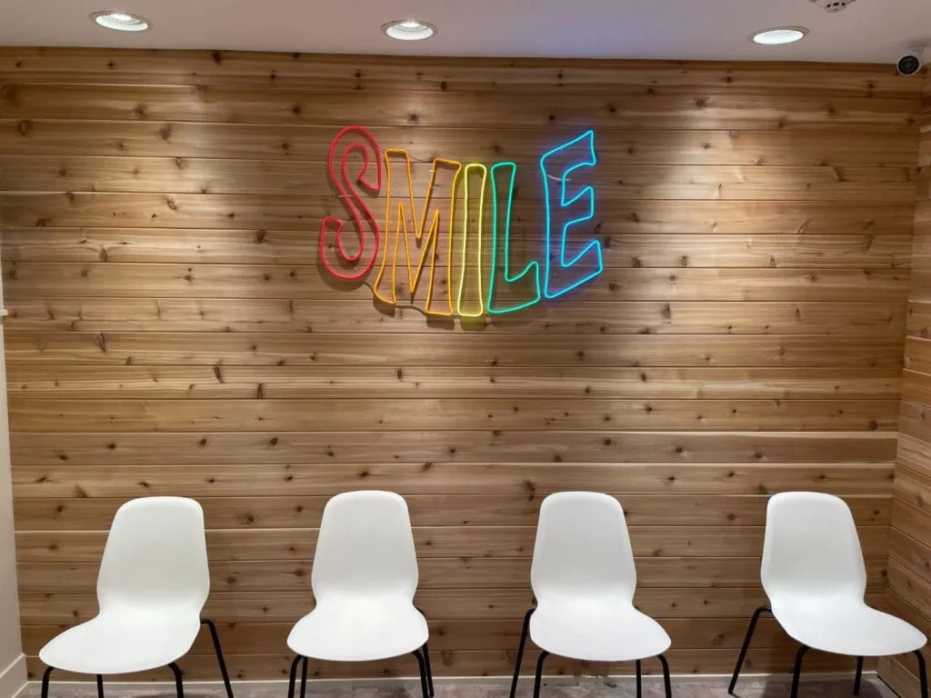 Brightly lit orthodontic waiting area featuring a wooden wall with a neon "SMILE" sign and four white chairs, reflecting the welcoming environment of Smile Orthodontics by Dr. Sarah.